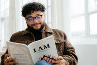 Man reading a book titled 'I AM: The Identity of Jesus' in a bright room with large windows.